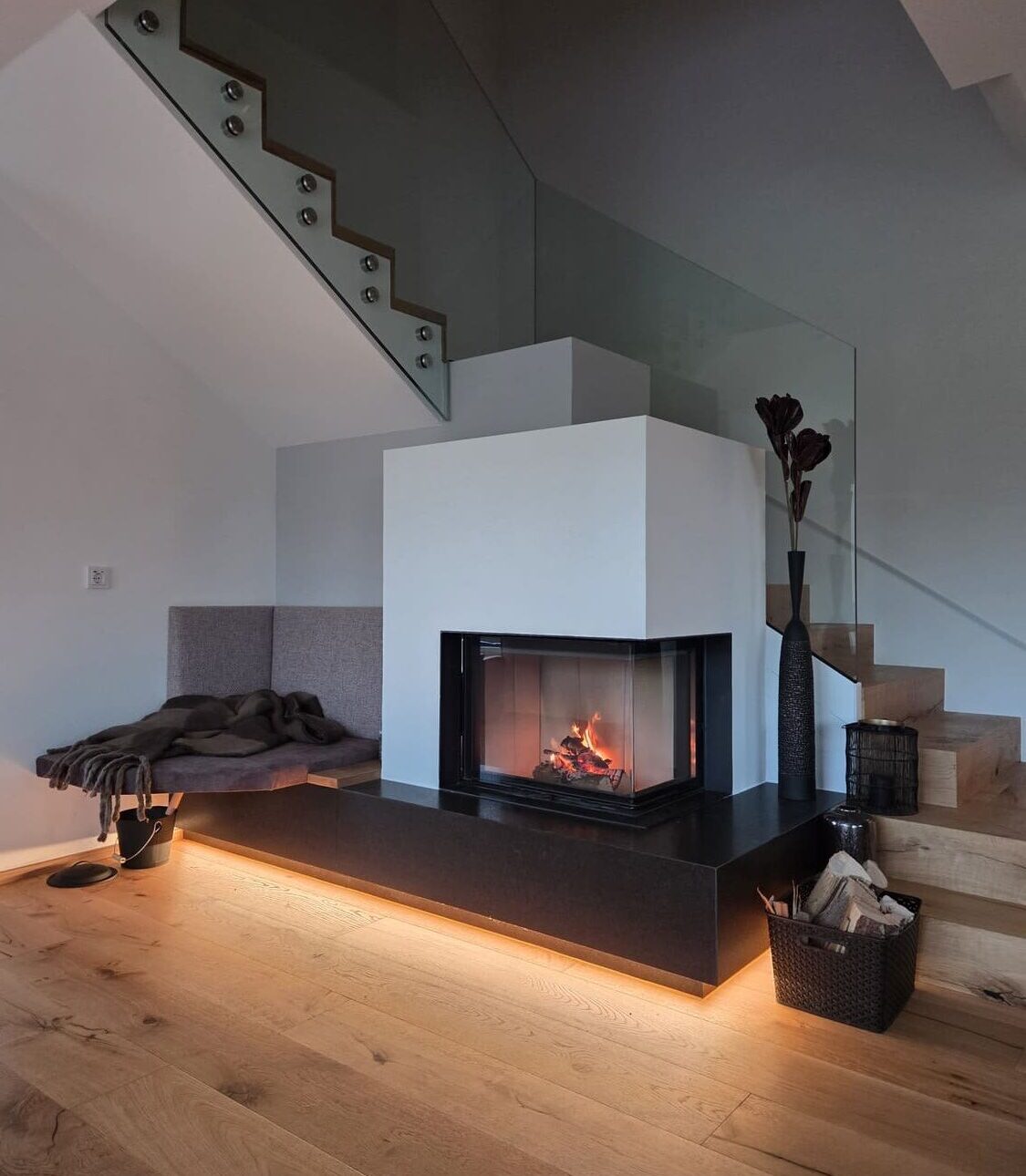 Moderner Kachelofen perfekt integriert in einer weißen Wand, der Boden ist aus Holz und dahinter befindet sich eine Stiege die in den ersten Stock führt