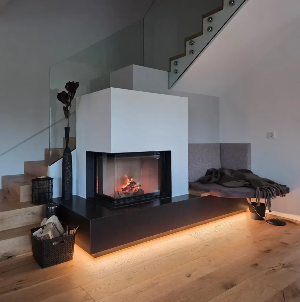 Moderner Kachelofen perfekt integriert in einer weißen Wand, der Boden ist aus Holz und dahinter befindet sich eine Stiege die in den ersten Stock führt