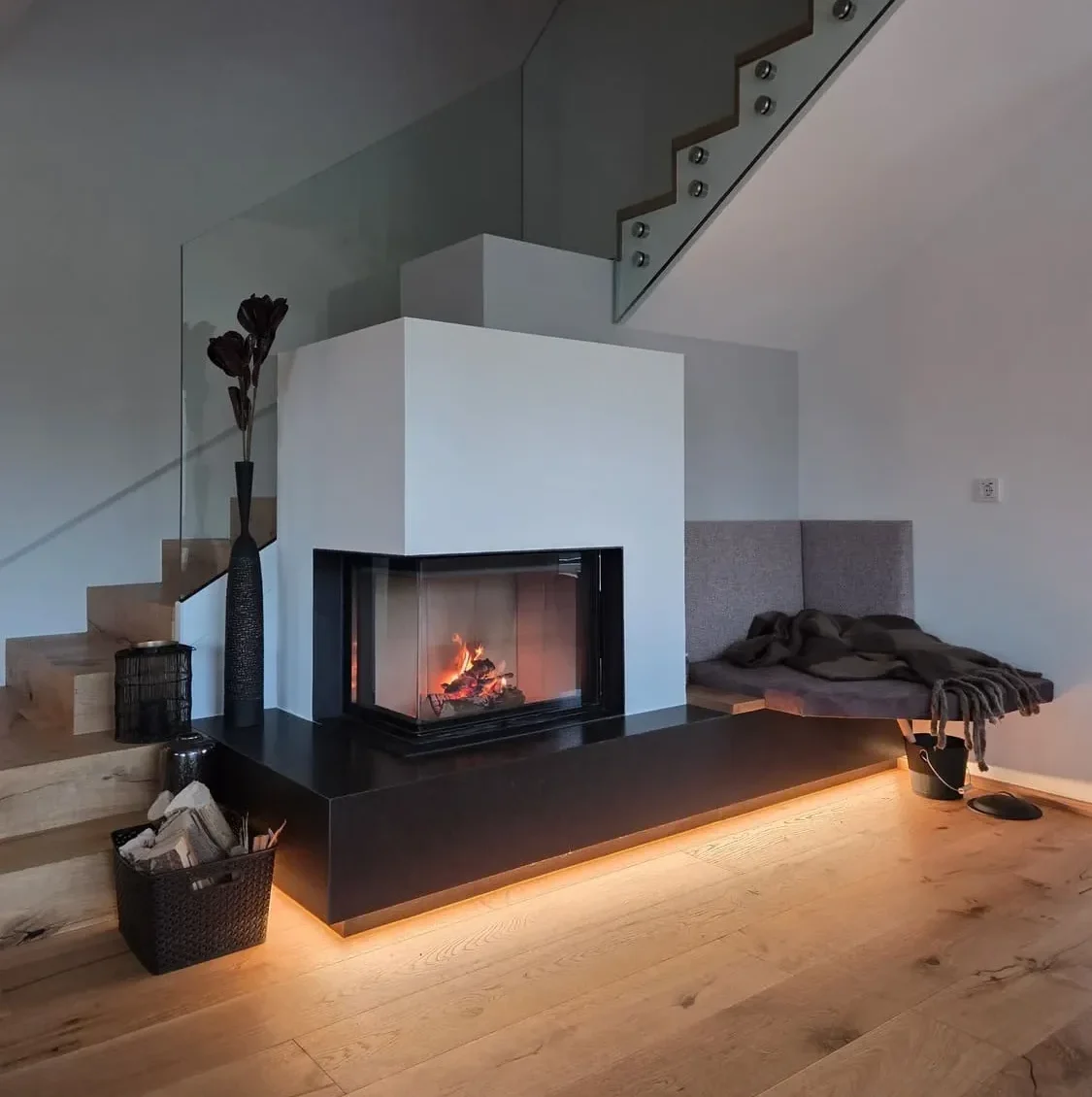 Moderner Kachelofen perfekt integriert in einer weißen Wand, der Boden ist aus Holz und dahinter befindet sich eine Stiege die in den ersten Stock führt